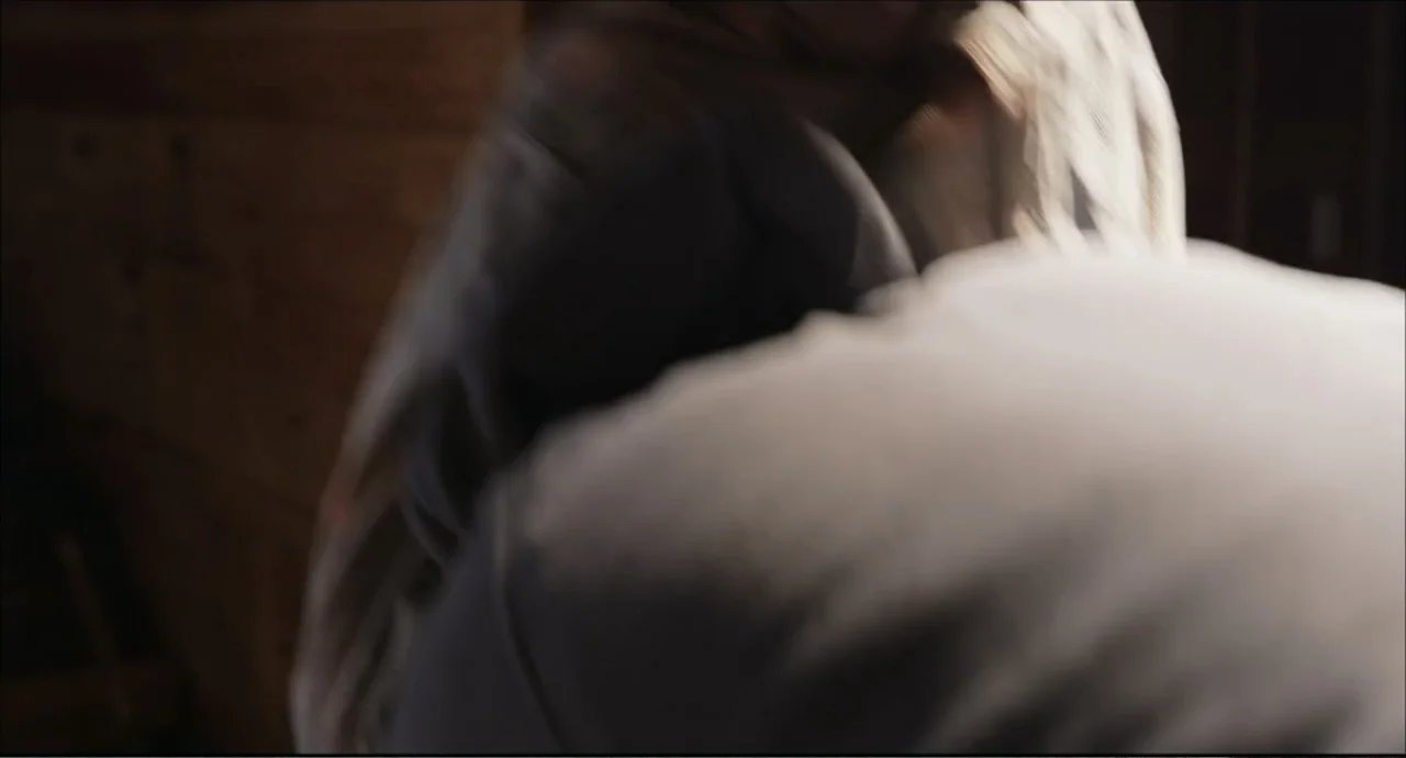 Freedom (2024) movie still: closeup — A dimly lit interior space features dark wood paneling on the side walls. Two individuals stand close together…