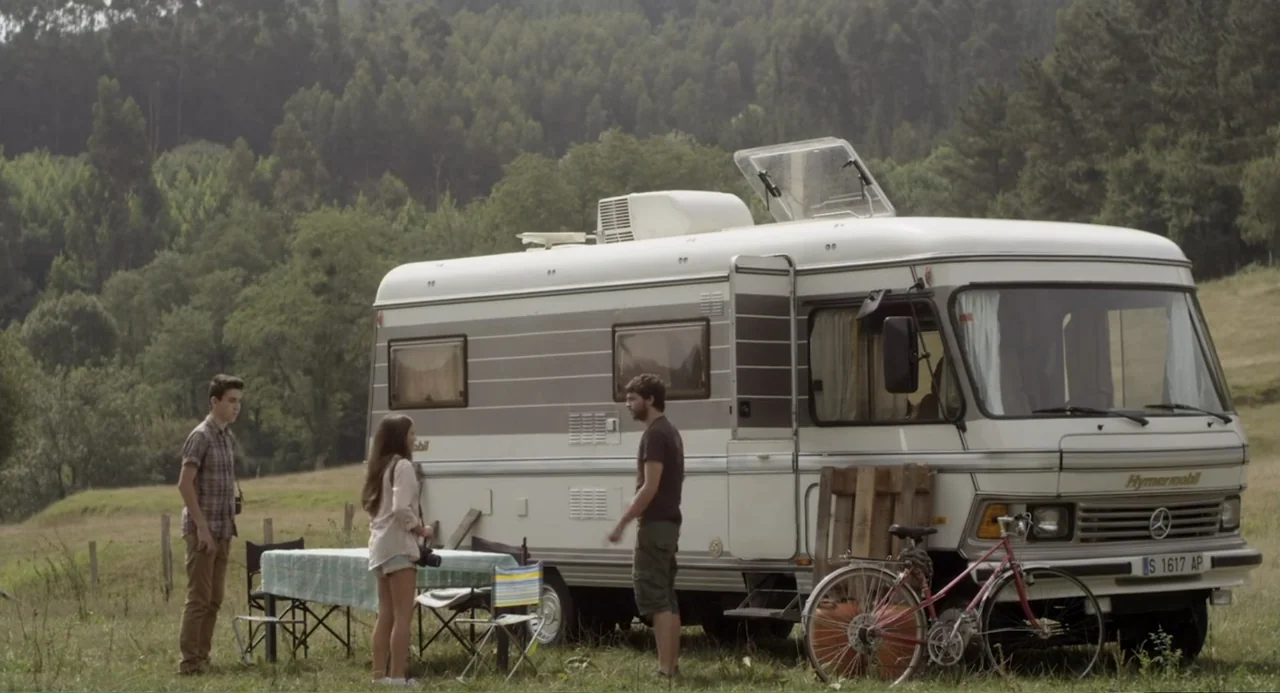 Newton's Third Law (2017) movie still: wide — A grassy meadow with a parked recreational vehicle near a dense tree line. Three people stand around a…