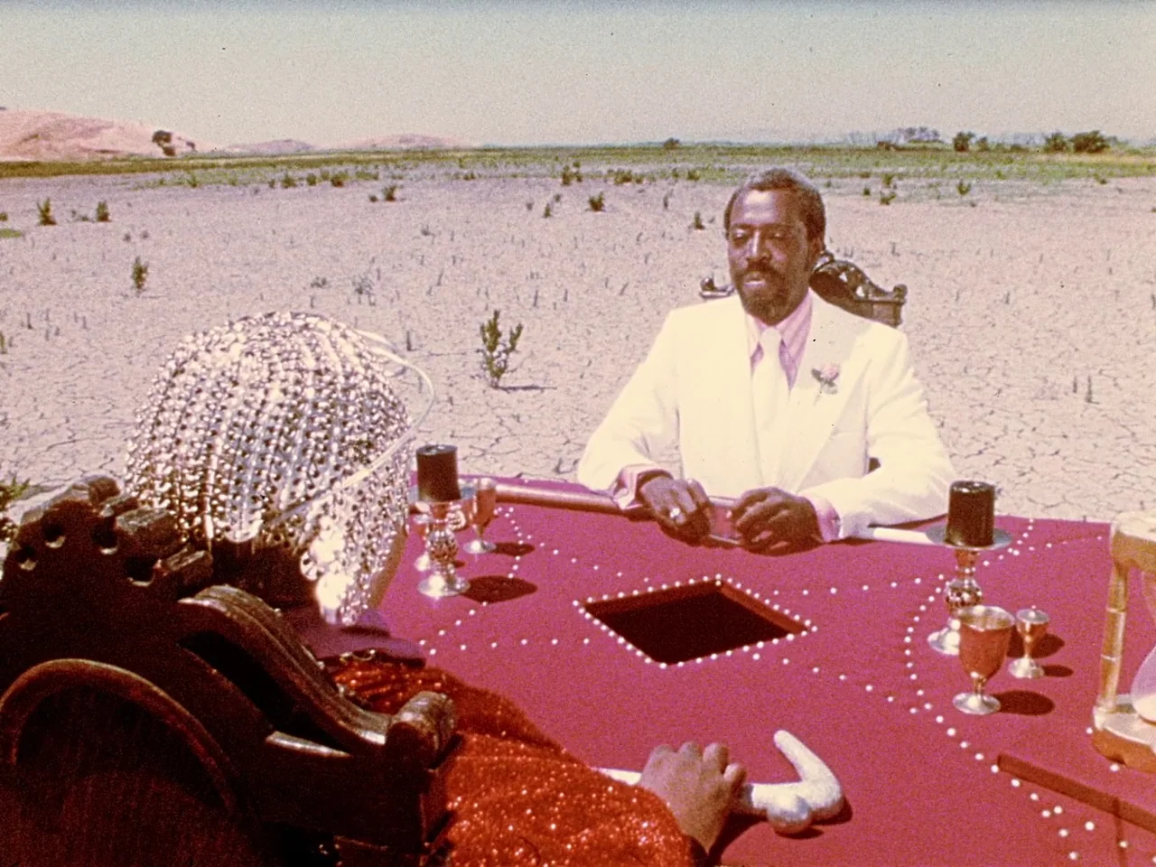 Space is the Place (1974) movie still: medium — A vast, arid dry lake bed under a clear, bright sky. Two people face one another across a…