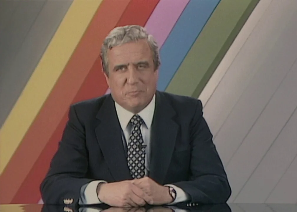 No (2012) movie still: medium — A television news studio features a backdrop with diagonal multicolored stripes. A middle-aged man in a suit sits at…