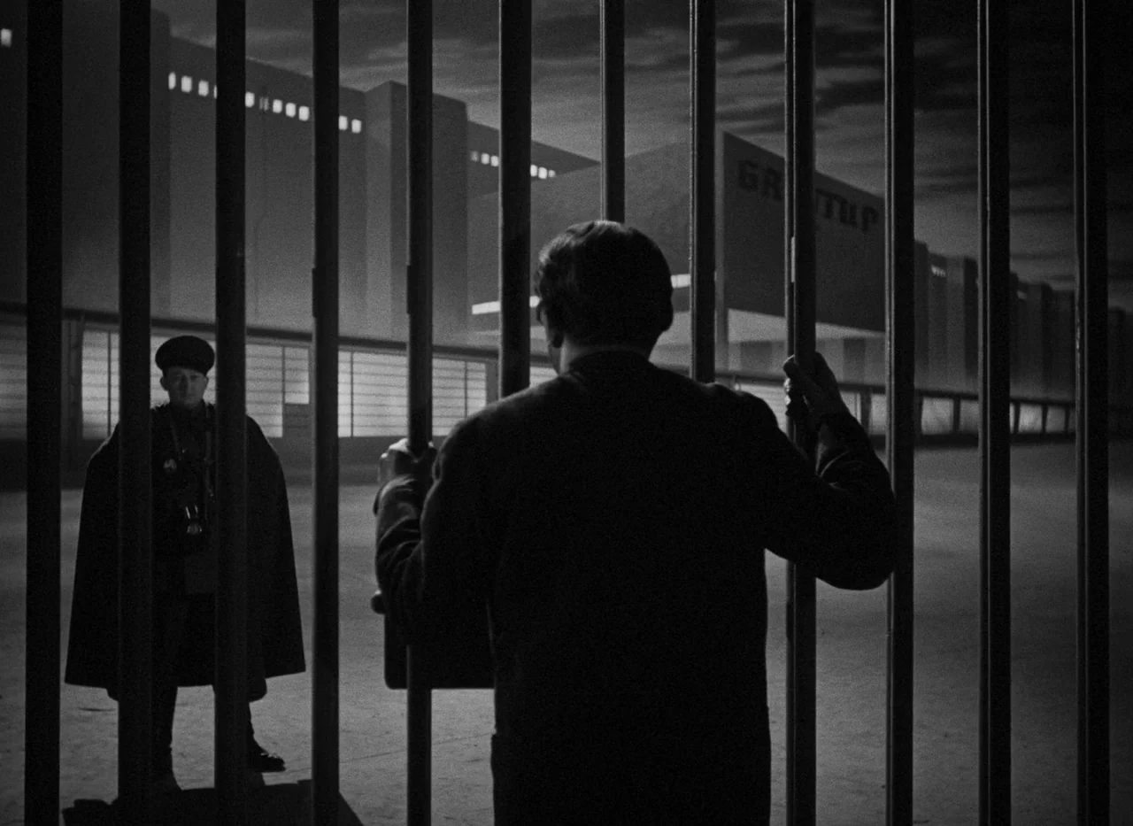 Krakatit (1948) movie still: medium — A dark night exterior features a man held behind heavy vertical metal bars. A guard in a military uniform and…