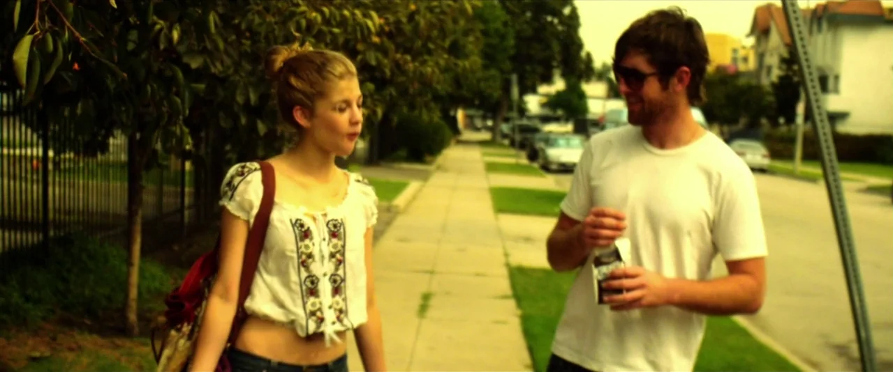 Bellflower (2011) movie still: medium — Two young adults stand on a concrete sidewalk beside a leafy tree. They walk together while conversing on a…