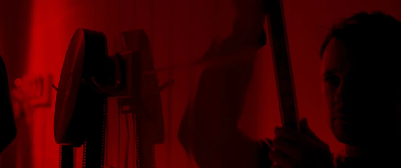 The Canal (2014) movie still: medium — A darkroom bathed in monochromatic red light containing film processing equipment. A man holds a strip of…