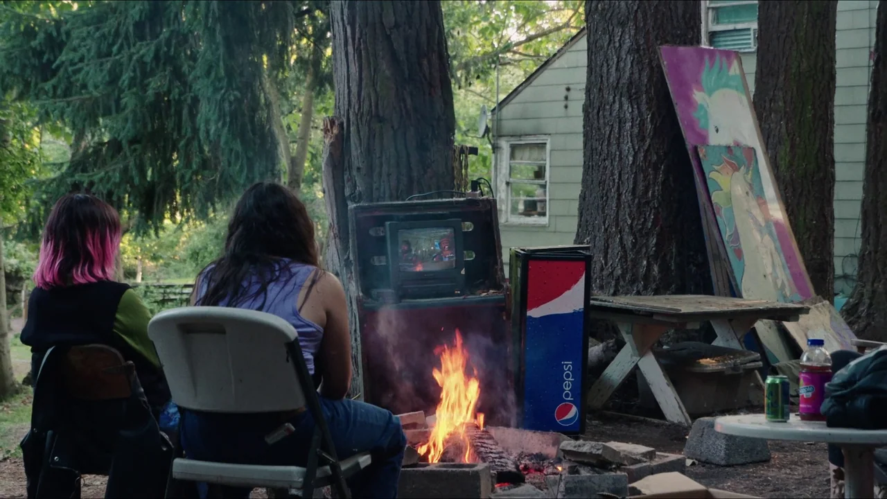 Gasoline Rainbow (2023) movie still: medium — A backyard setting features two people sitting on plastic chairs before a small campfire. A portable…