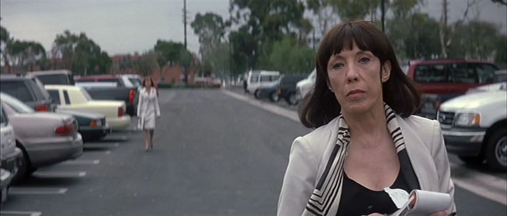 I Heart Huckabees (2004) movie still: medium — A paved asphalt parking lot filled with various cars on a cloudy day. A woman in a light jacket walks…