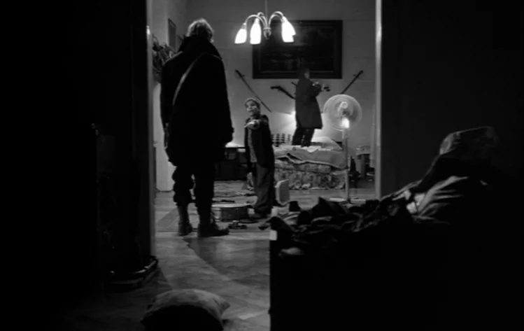 Werckmeister Harmonies (2000) movie still: wide — A dark, cluttered apartment room features a bed, a pedestal fan, and a chandelier. A tall person…