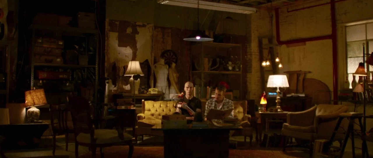 Depraved (2019) movie still: wide — A cluttered, dimly lit basement room features exposed brick walls and scattered vintage furniture. Two men sit…