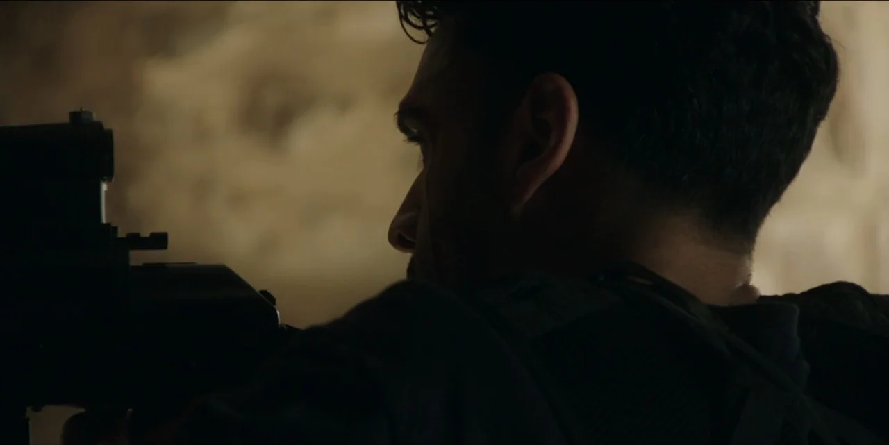 The Hunted (2024) movie still: closeup — A dark indoor space with textured wall surfaces visible in the background. A man in tactical gear aims a…