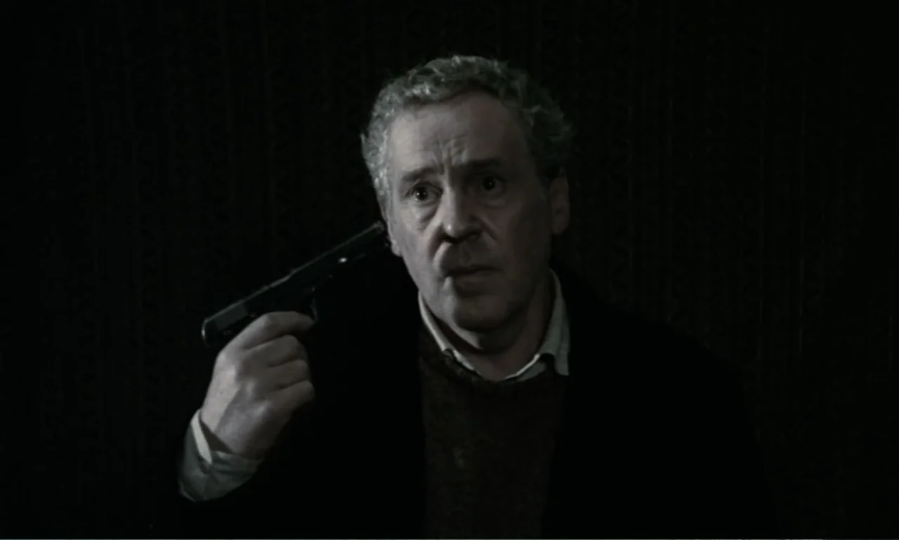 The Sacrifice (2015) movie still: medium — A dimly lit interior space features a man standing against a dark, textured wall. The man holds a black…