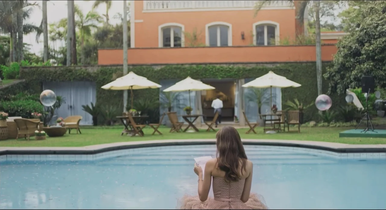 Proof of Love (2022) movie still: medium — A terracotta-colored villa stands behind a manicured lawn with tables and patio umbrellas. A young woman…