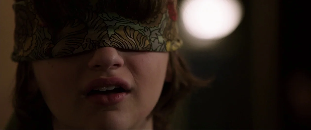 The Conjuring (2013) movie still: closeup — A close-up shot of a person wearing a patterned fabric blindfold over their eyes. The background is dark…