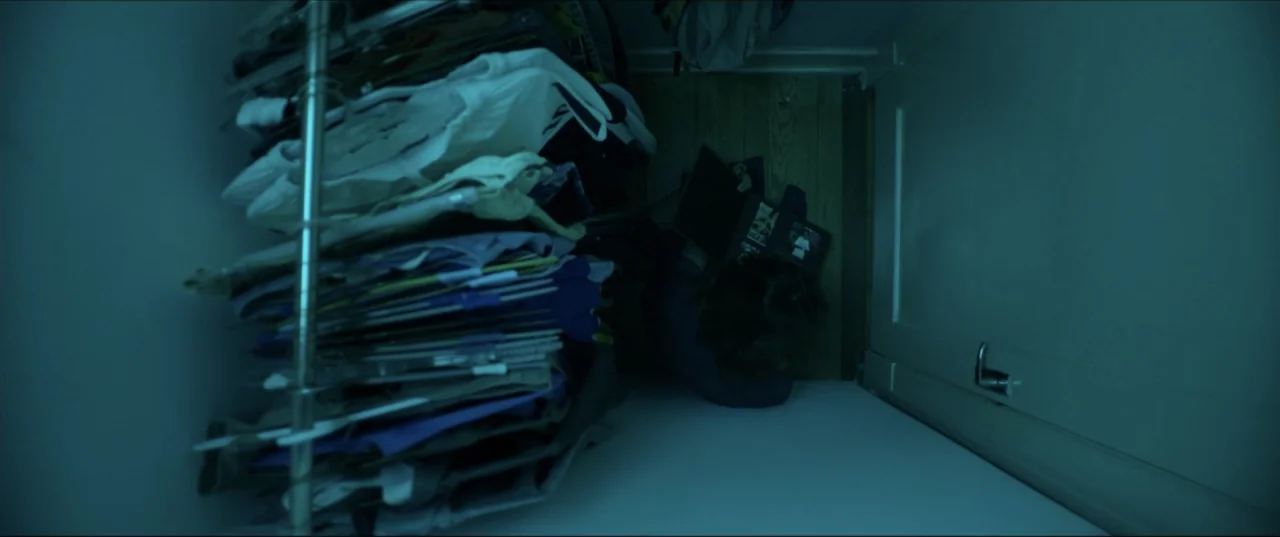 Can't Quit Mom (2022) movie still: wide — A dark, cramped closet with a metal clothing rack stacked with numerous folded garments. A person is…