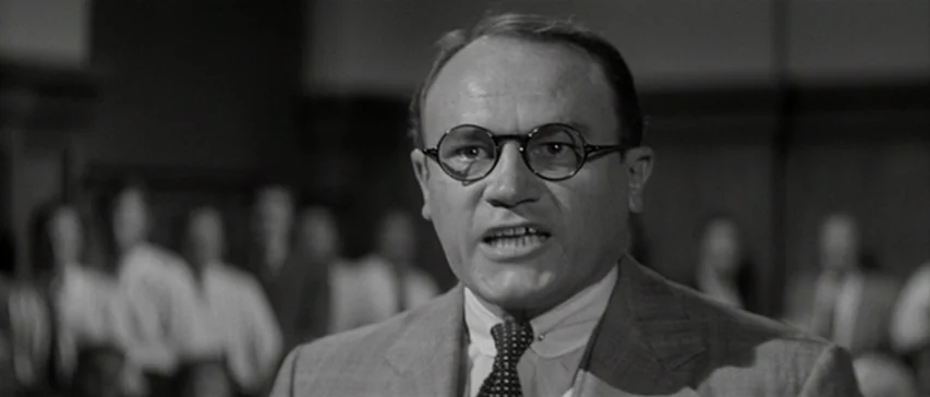 Compulsion (1959) movie still: closeup — A courtroom with blurred figures in the background seated in rows. A man in glasses speaks directly toward…
