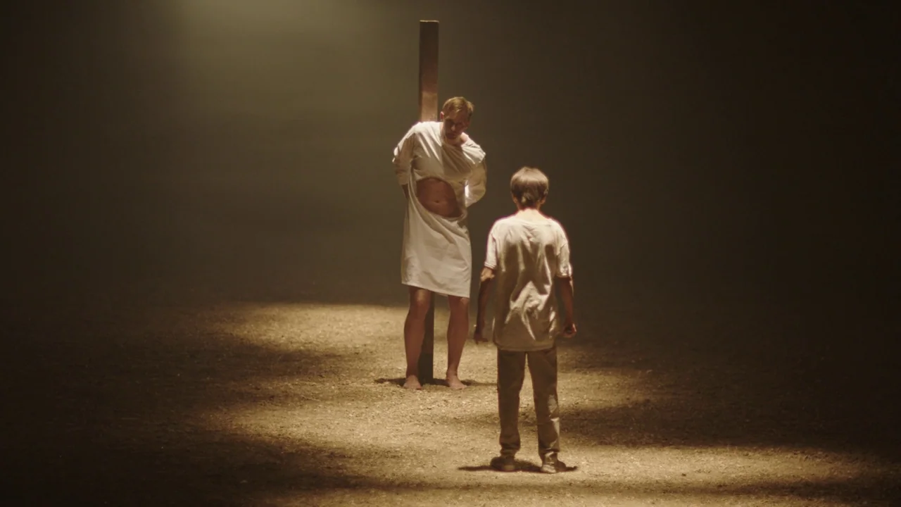 Infinity Pool (2023) movie still: fullbody — A dark, dusty outdoor space features a lone vertical wooden post in the center. An adult male stands…