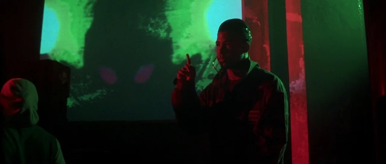 Bones movie still: medium — A dimly lit industrial room illuminated by a large screen playing bright green footage. A man stands in the foreground…
