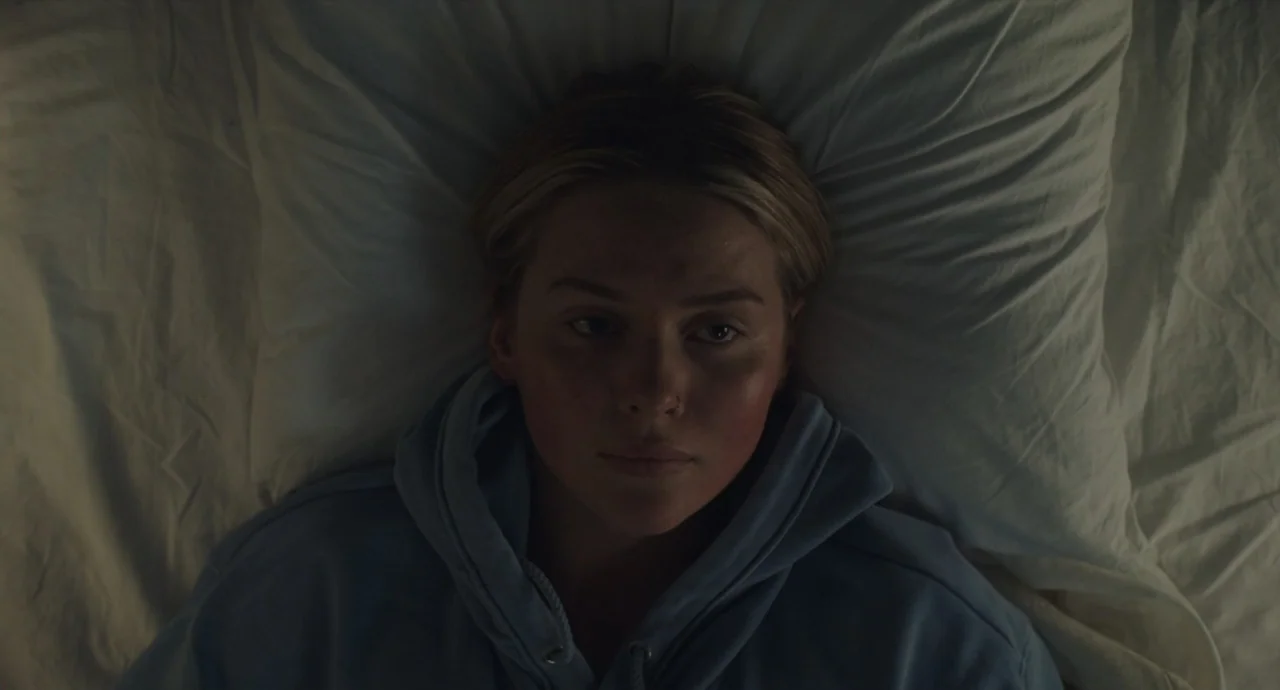Pleasure (2021) movie still: closeup — A person lies on white bedsheets within a dimly lit space. The subject faces the camera directly while…