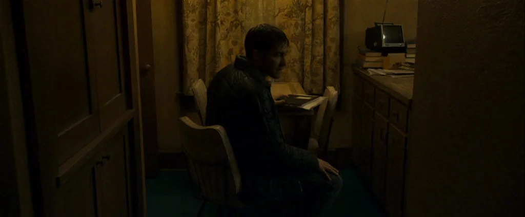 Zodiac (2007) movie still: medium — A dimly lit room contains a wooden table and chairs with floral patterned curtains. A man sits alone at the…