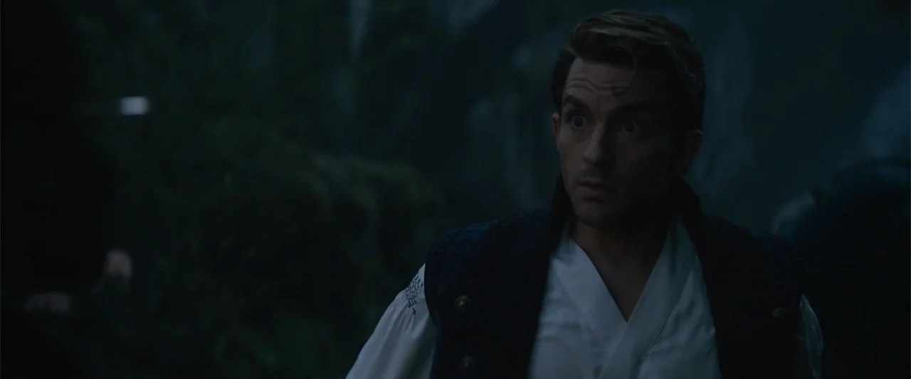 Wicked (2024) movie still: medium — A dark, out-of-focus outdoor area with dense foliage and low ambient light. A man in period-appropriate attire…