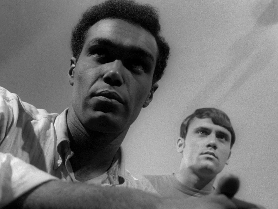 Night of the Living Dead (1968) movie still: medium — A plain indoor setting serves as the backdrop for two men. The subjects observe something…