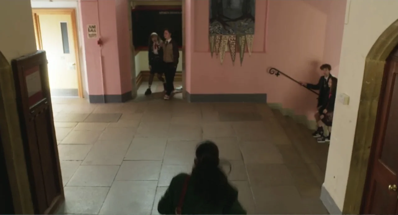 How to Date Billy Walsh (2024) movie still: medium — A school interior features pink walls, stone flooring, and several students standing together.…