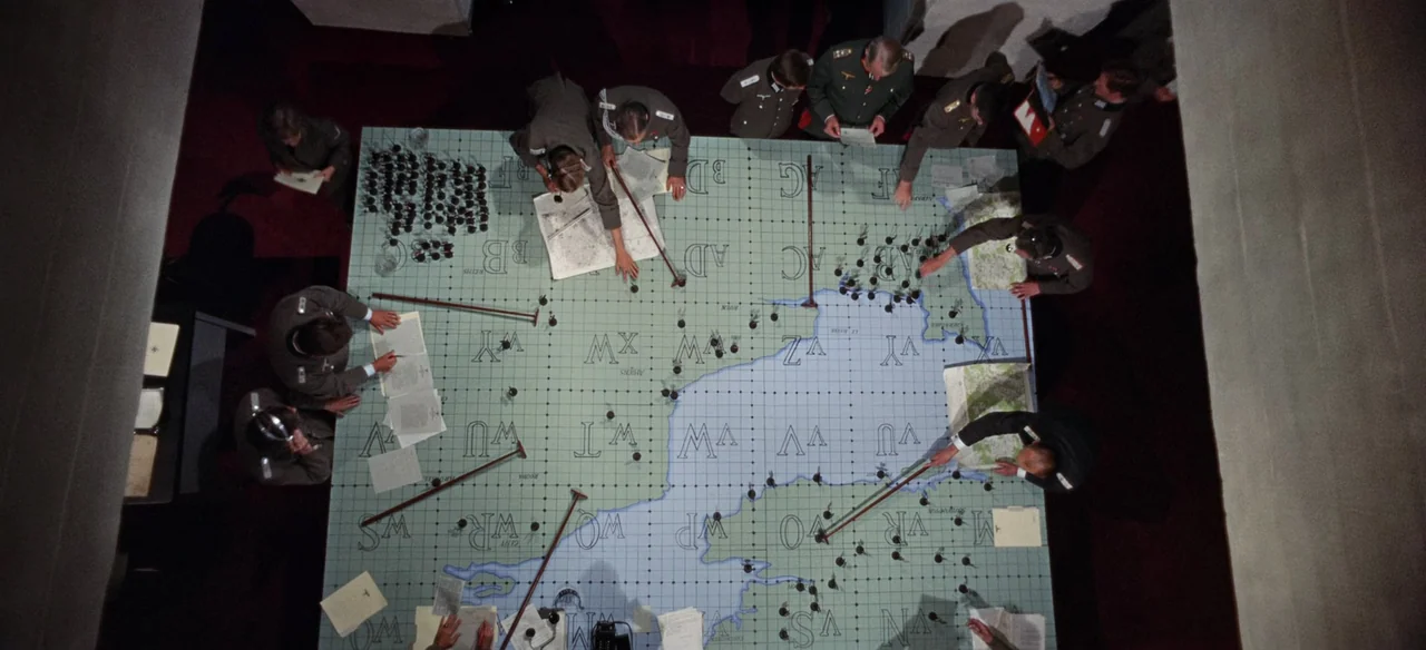 Patton (1970) movie still: wide — A high-angle view shows multiple military officers gathered around a large gridded map table. The men use long…