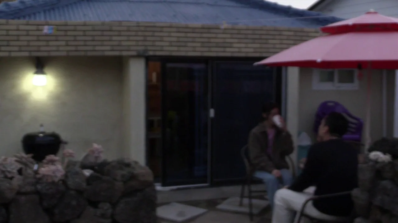 In Water (2023) movie still: wide — An outdoor patio area features a brick-walled structure with a lit wall-mounted fixture. Two men sit in metal…