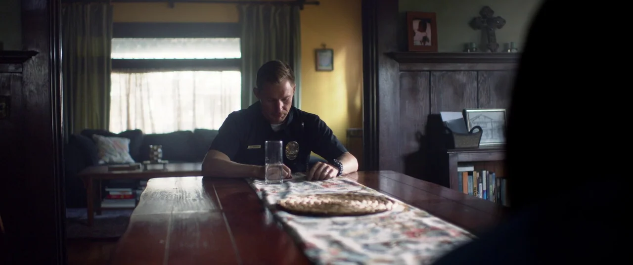 Blindfire (2020) movie still: medium — A residential dining area features a dark wood table and a fireplace with mantle decor. A police officer sits…