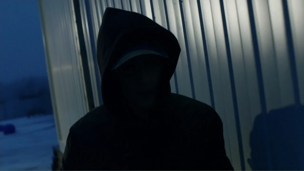 Code of Trust (2019) movie still: medium — An exterior industrial space features a corrugated metal wall against a dark night sky. A lone figure in…