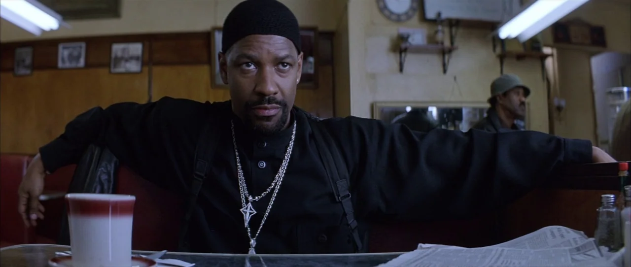 Training Day (2001) movie still: medium — A man sits at a booth in a dimly lit diner with wooden walls. The man rests his arms along the back of the…