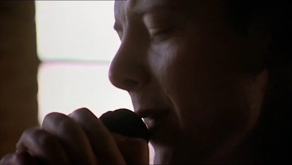 24 Hour Party People movie still: closeup — A close profile of a man singing into a handheld microphone inside a studio. Bright window light bleeds…