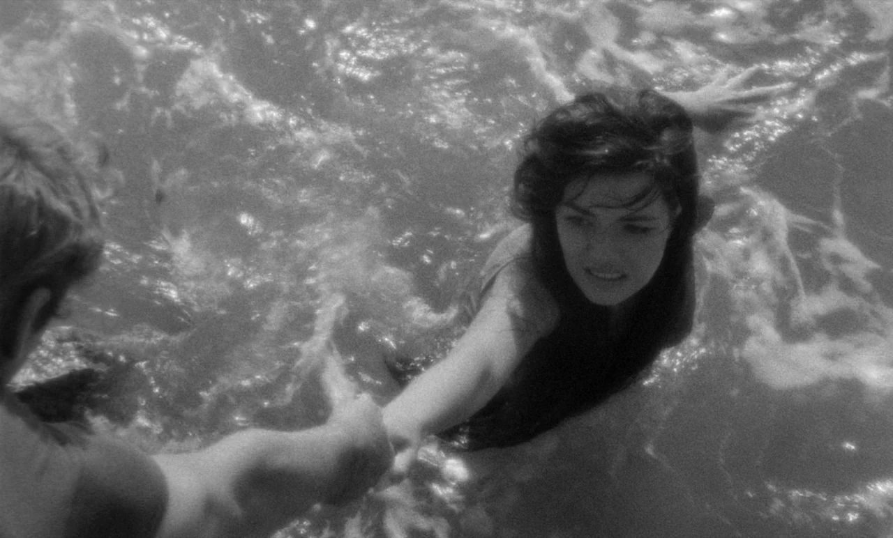 Night Tide (1961) movie still: medium — Two individuals are partially submerged in bright, choppy ocean water under direct sunlight. One person…