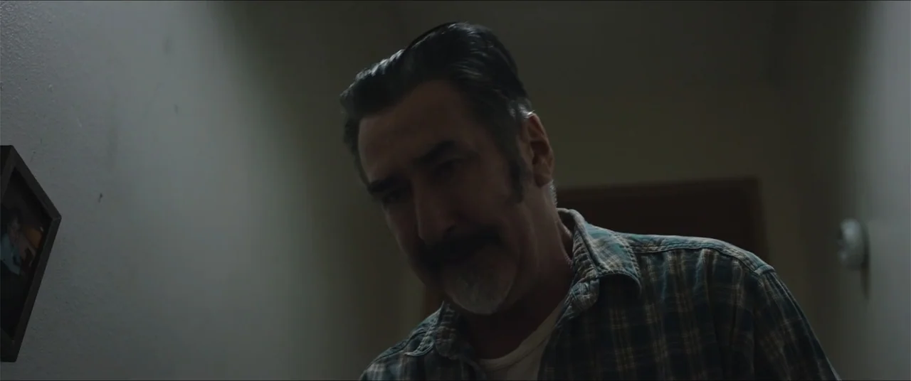 Captive (2020) movie still: medium — A dimly lit narrow residential hallway with a dark wall and a mounted picture frame. A middle-aged man with…