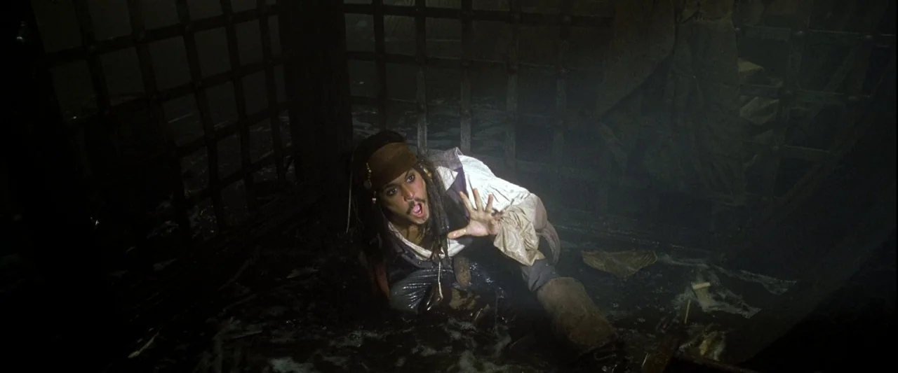Pirates of the Caribbean: The Curse of the Black Pearl movie still: medium — A dark, damp stone dungeon features iron bars and puddles of water on…