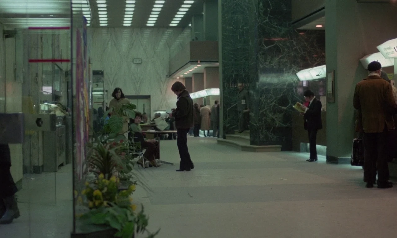 The Margin (1976) movie still: wide — A 1970s airport terminal hall features green marble pillars and recessed fluorescent lighting grids. People…