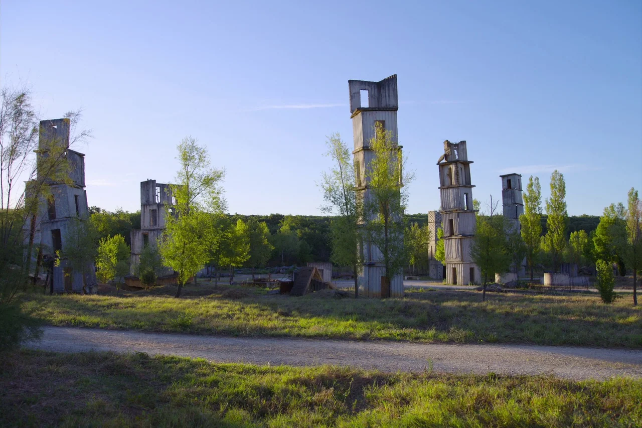 Anselm (2023) movie still: establishing — Several concrete skeletal tower structures are situated across a wide, grassy landscape. Green deciduous…