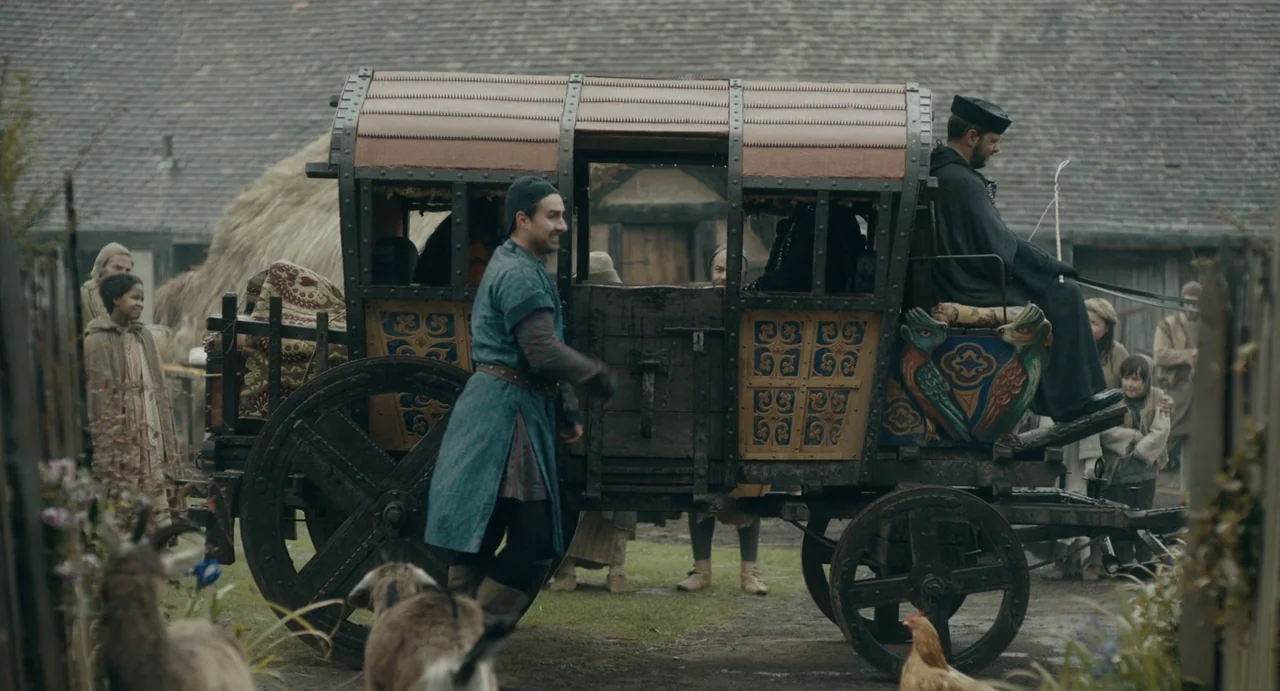 Catherine Called Birdy (2022) movie still: wide — A rural medieval village setting features a wooden wagon parked on a dirt path. A man in a blue…