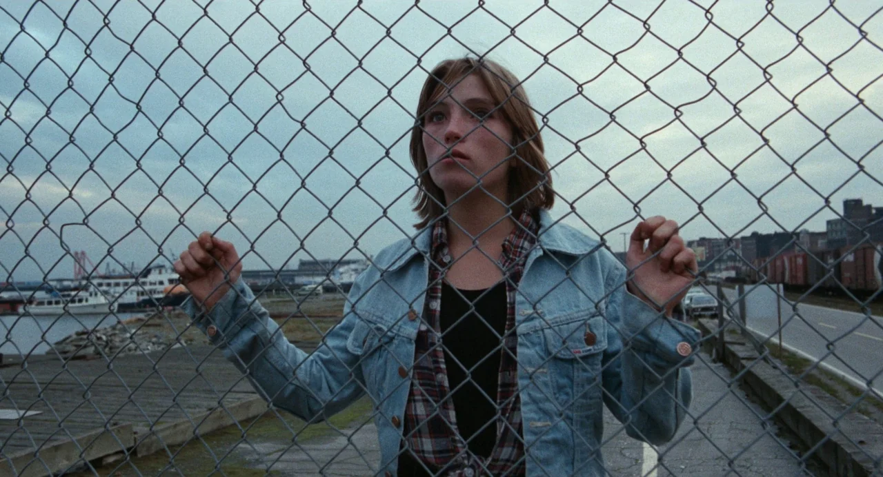 Out of the Blue (1980) movie still: medium — An outdoor industrial area features a rusted chain link fence obscuring a coastal view. A young woman…