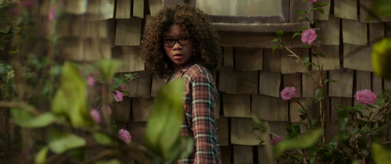 A Wrinkle In Time (2018) movie still: medium — A wooden exterior wall with weathered shingles serves as the backdrop. The girl stands amidst a patch…