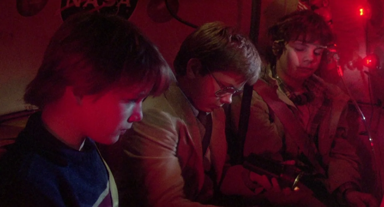 Explorers (1985) movie still: medium — Three teenage boys sit closely together in a dim, red-lit craft with NASA branding. The group examines small…