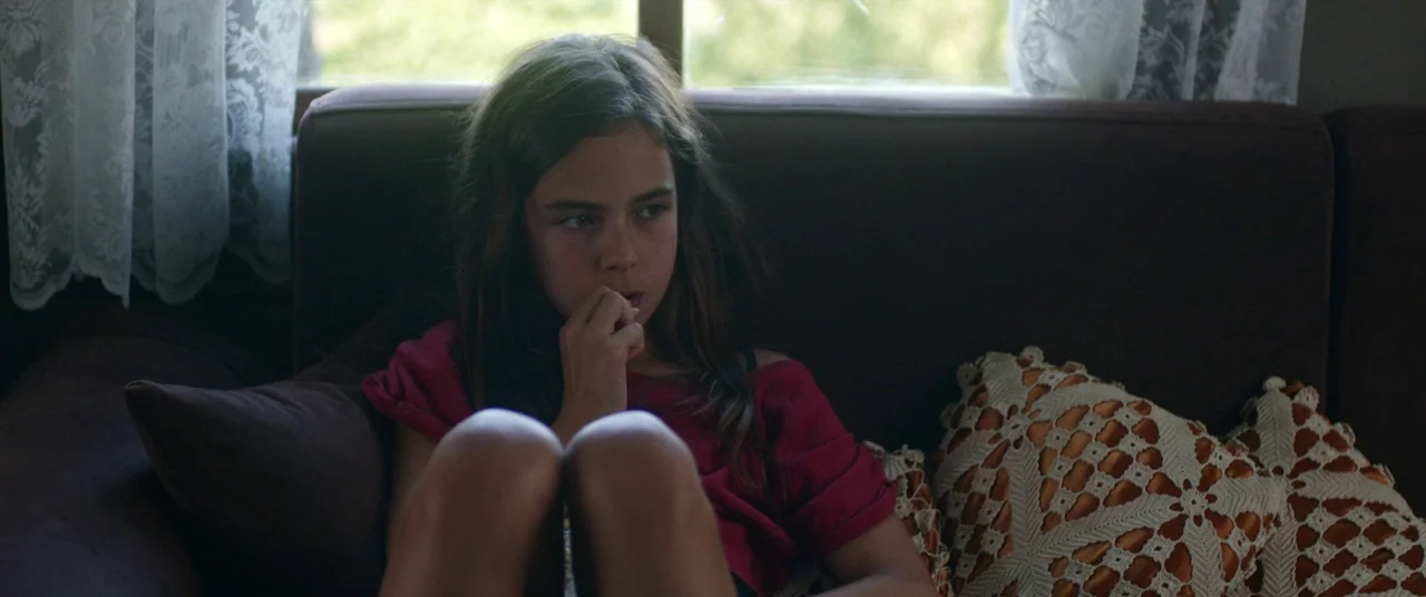 Mustang (2015) movie still: medium — A living room interior featuring a brown fabric sofa decorated with crochet pillows. A young girl sits on the…