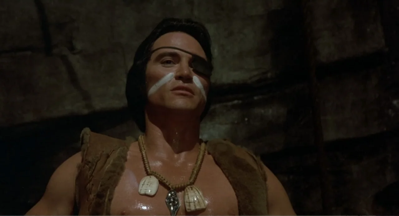 Firewalker (1986) movie still: closeup — A man stands inside a dark, rough-hewn stone chamber illuminated by flickering light. He gazes upward,…