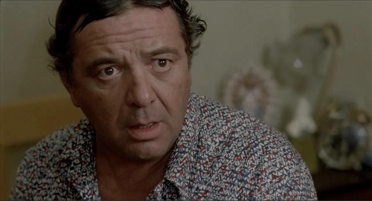 Waiting for the Hearse (1985) movie still: closeup — A domestic interior with a man in the foreground and blurred decor behind him. The man is…