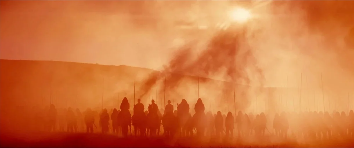 Macbeth (2015) movie still: wide — A vast, flat landscape is filled with a large, motionless army of people and horses. Thick orange dust and heavy…