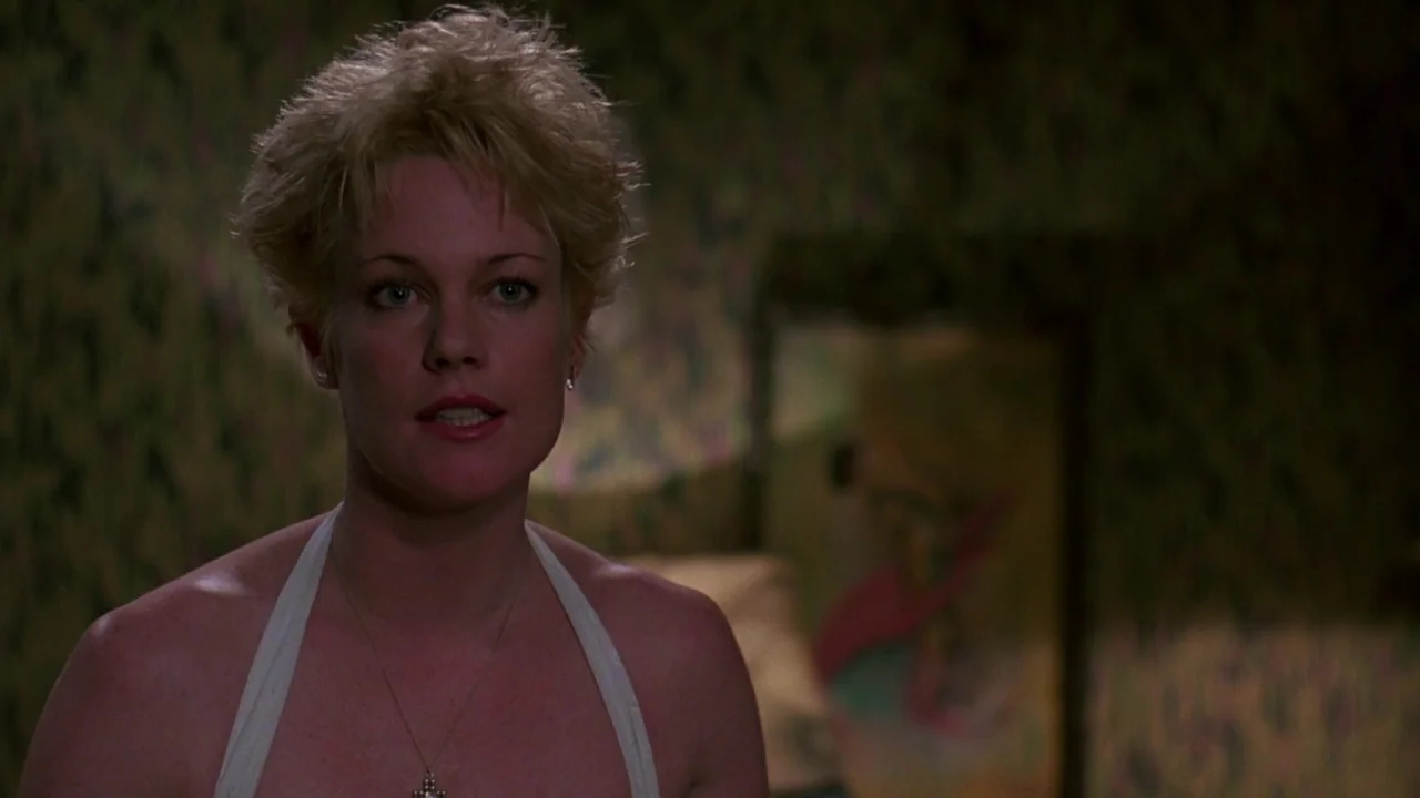 Something Wild (1986) movie still: closeup — A dimly lit room features patterned wallpaper and a blurred artwork on a wall. A woman with short…