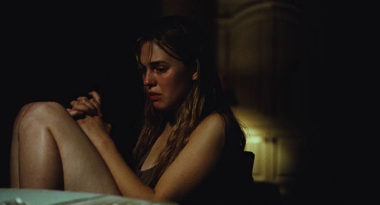 The Giant (2019) movie still: medium — A dimly lit room with a single warm light illuminating the side of a face. A woman sits at a table with her…