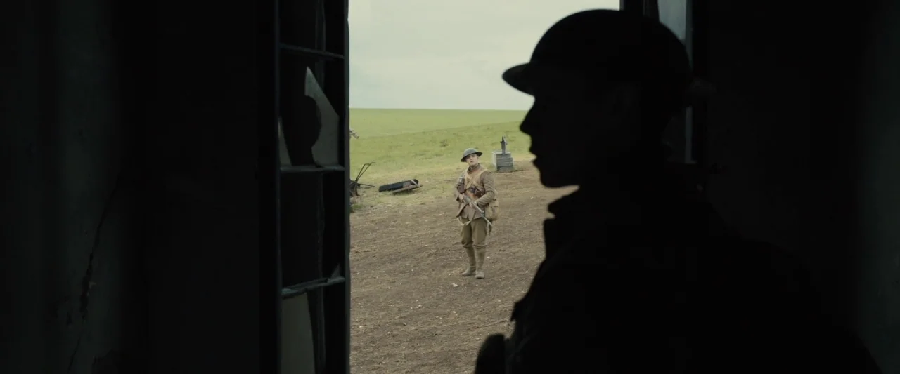 1917 movie still: wide — An open dirt field with green grass under an overcast sky viewed from a doorway. A soldier in uniform stands in the…