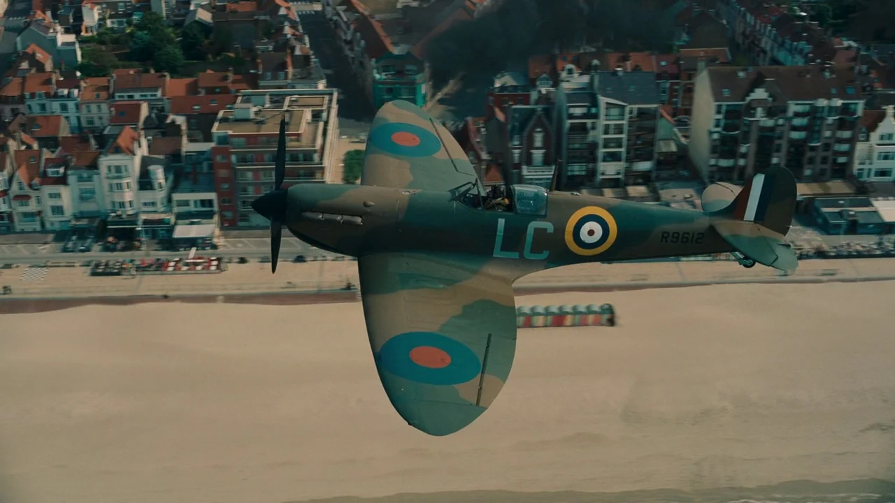 Dunkirk (2017) movie still: wide — A green-camouflaged Spitfire flies low over a sandy beach near a coastal town. The fighter plane banks slightly…