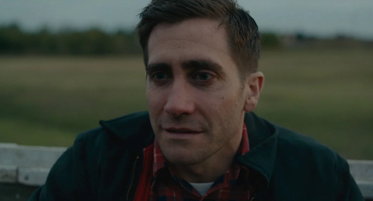 Wildlife (2018) movie still: medium — An open field under a flat, overcast sky during twilight hours. A man with short dark hair gazes off-frame…