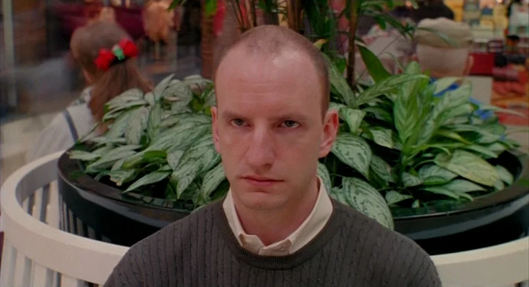 Schizopolis movie still: closeup — A man sits in front of a circular planter inside a shopping mall corridor. The mall background is partially…