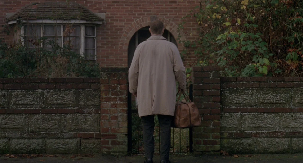 Possum (2018) movie still: fullbody — A brick residence with an arched entryway is partially obscured by overgrown bushes. A man holding a leather…